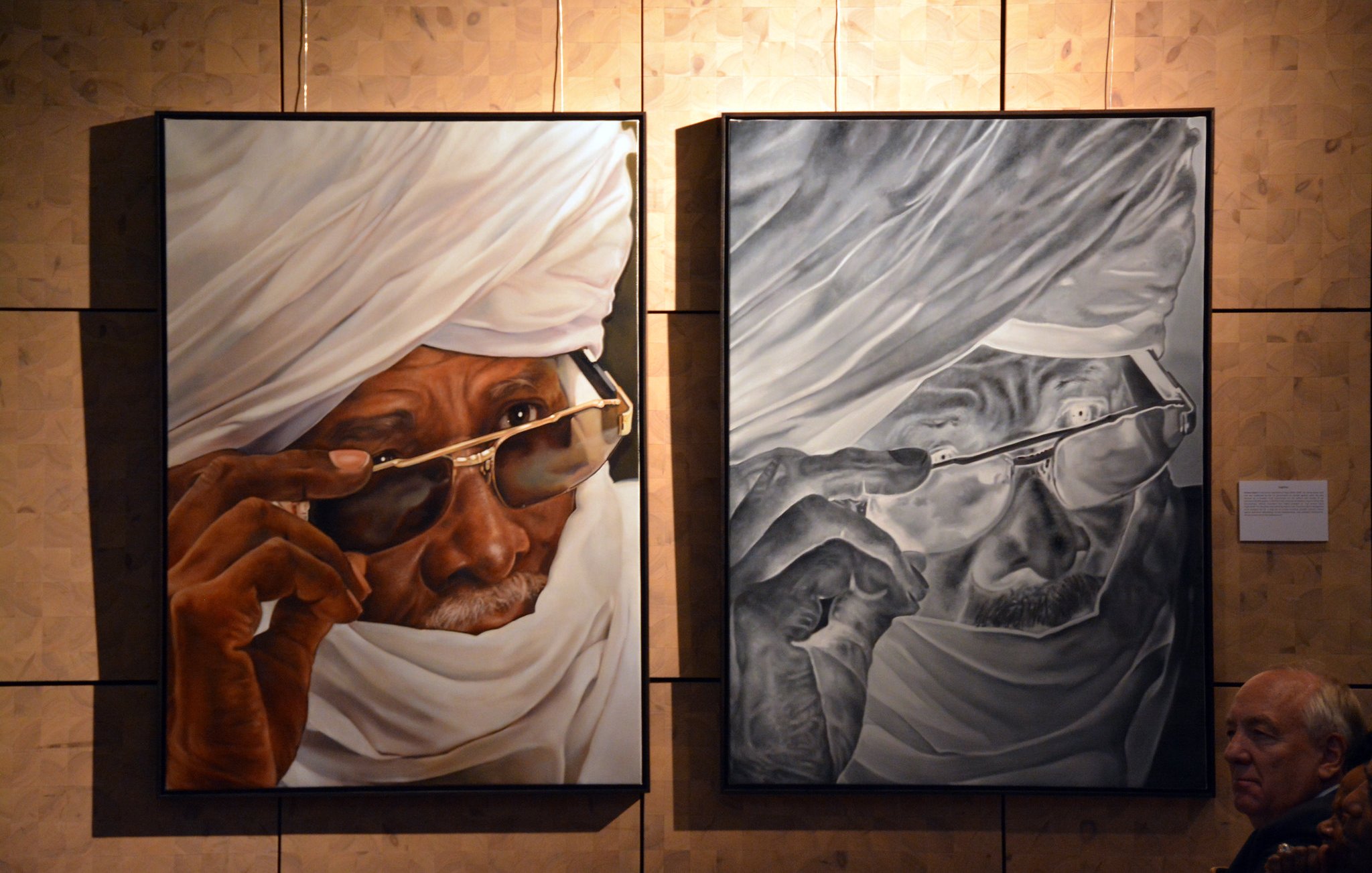 L'image présente deux tableaux accrochés sur un mur, représentant le même homme, mais dans des styles différents. À gauche, le portrait est en couleurs vives : un vieil homme porte un turban blanc et des lunettes de soleil. Son regard est pensif, avec une expression de sagesse. À droite, l'œuvre est en noir et blanc, offrant un contraste fort. Les détails des rides et des textures de la peau sont accentués, donnant une sensation de profondeur. En bas à droite de l'image, on aperçoit une silhouette d'un visiteur qui observe les œuvres. Le fond du mur est constitué de panneaux en bois clair, ajoutant une dimension chaleureuse à l'espace d'exposition.