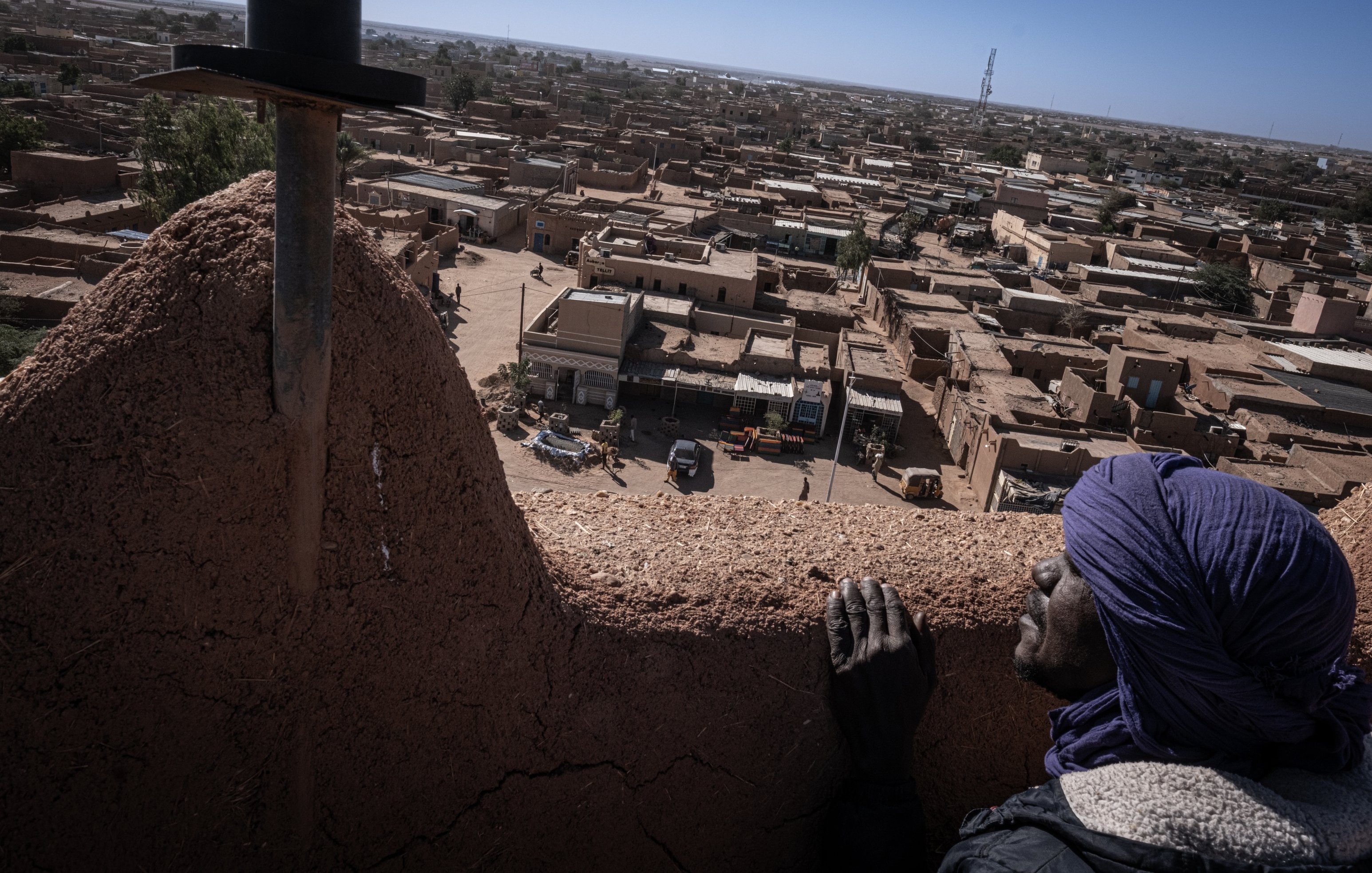 L'image présente une vue panoramique d'une ville saharienne. Au premier plan, une personne au visage marqué par le temps s'appuie sur un mur en terre. Elle porte un turban et semble contempler le paysage. En arrière-plan, des maisons en adobe s'étendent à perte de vue, avec des toits plats et des teintes ocres. On peut apercevoir des petites routes et des véhicules qui circulent. L'horizon est dégagé, avec quelques structures modernes qui contrastent avec l'architecture traditionnelle. L'atmosphère évoque à la fois la chaleur du désert et la tranquillité d'une ville endormie.
