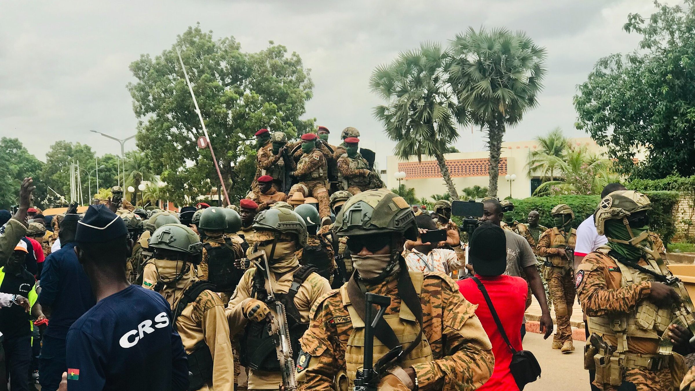 L'image montre une scène animée en plein air, probablement lors d'un événement public ou d'une manifestation. Au premier plan, plusieurs soldats sont regroupés, portant des uniformes militaires et des casques. Ils sont armés et semblent vigilants. L'un d'eux est sur un véhicule militaire, tandis que d'autres avancent sur la route. En arrière-plan, des arbres et des bâtiments peuvent être aperçus, et une foule de citoyens se trouve à proximité, certains portant des vêtements colorés. L'atmosphère peut sembler tendue, surtout en raison de la présence militaire. Le ciel est nuageux, ajoutant une certaine gravité à la scène.