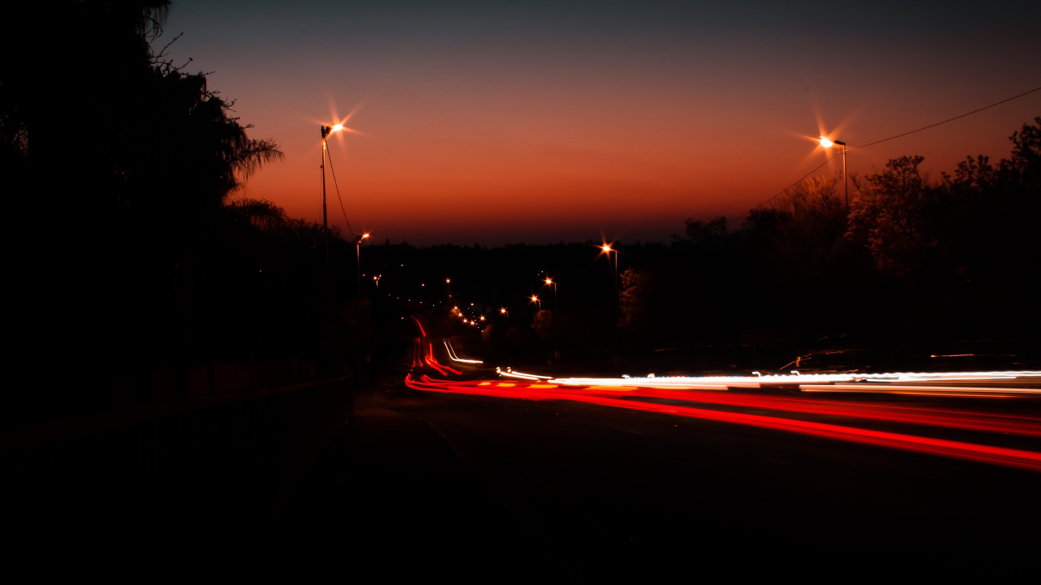 Imagine une scène tranquille à l'heure où le jour se transforme en nuit. Le ciel se teinte de dégradés d'orange et de bleu sombre, créant une ambiance douce et apaisante. Au loin, on aperçoit une route bordée d'arbres, presque silhouettes noires se découpant à l'horizon. Des lampadaires illuminent le chemin, leurs lumières jaunies ajoutant une touche chaleureuse à la scène. Des traces de lumière, comme des lignes fluides et vives, suggèrent des voitures qui passent rapidement, laissant derrière elles des filaments rouges et blancs. L'ensemble crée une atmosphère calme, invitant à la contemplation de ce moment transitionnel entre le jour et la nuit.