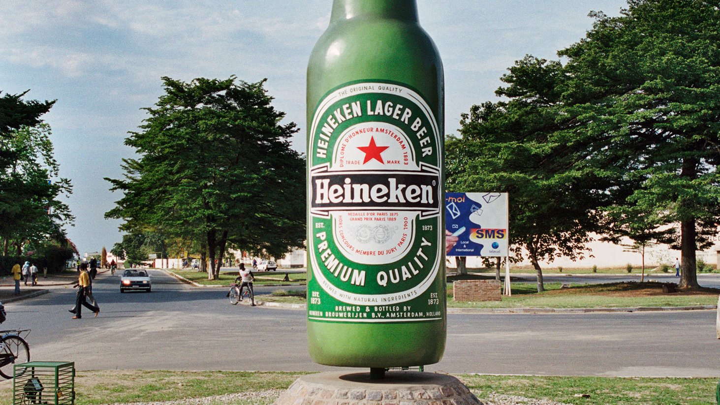 L'image montre une grande sculpture de bouteille de bière Heineken, qui se dresse au centre d'une intersection. La bouteille est peinte en vert vif et arborant l'étiquette rouge et blanche emblématique de la marque. Autour de la sculpture, on peut voir des gens se déplacer, certains à pied et d'autres à vélo. En arrière-plan, on distingue des arbres et quelques bâtiments qui ajoutent une ambiance locale. L'ensemble dégage une atmosphère animée et colorée, typique de l'urbanisme d'une ville vivante.