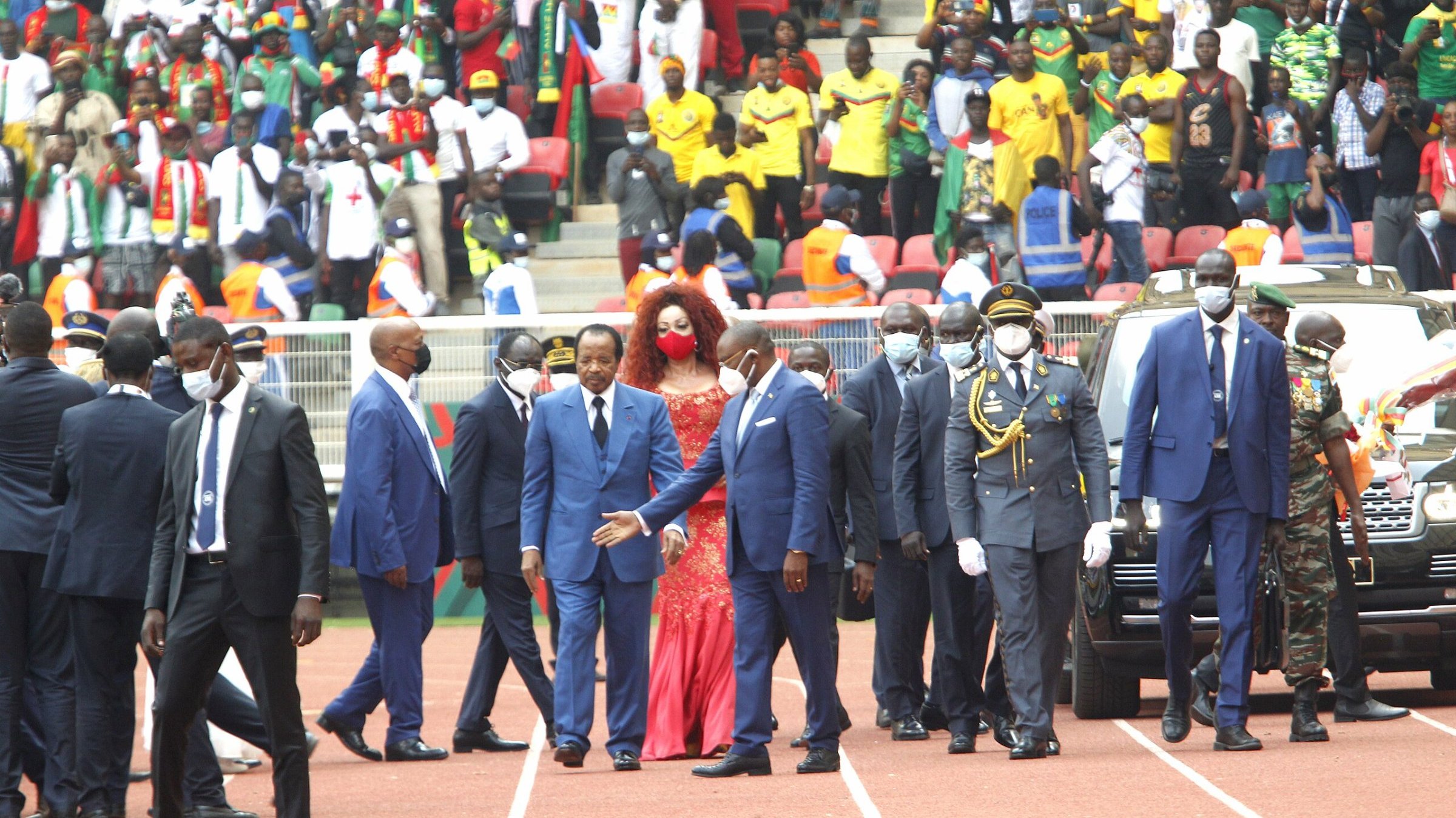 L'image montre un événement en extérieur, probablement un stade rempli de spectateurs. Au premier plan, un groupe de personnalités, dont plusieurs hommes en costume sombre et cravate, marche avec assurance. Parmi eux, une femme se distingue grâce à sa robe rouge éclatante et sa coiffure volumineuse. Ils semblent être en route pour un moment important, entourés de sécurité. À l'arrière-plan, on peut deviner une foule colorée et animée, avec des supporters portant divers vêtements aux couleurs vives, témoignant d'une atmosphère festive. L'ambiance générale semble être celle d'une grande célébration ou d'une cérémonie officielle.