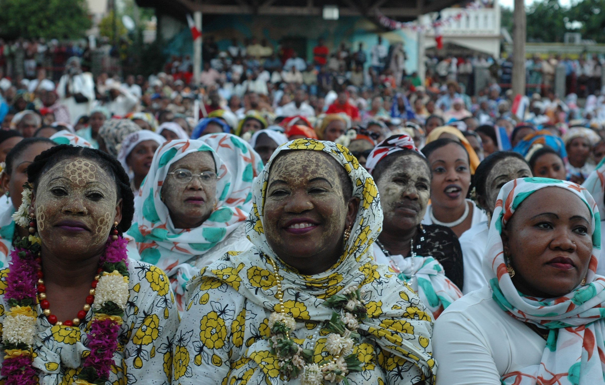 L'image montre un large groupe de personnes réunies pour un événement, probablement festif. Au premier plan, des femmes portent des vêtements colorés et traditionnels, agrémentés de motifs floraux. Certaines ont le visage recouvert de farine ou d'une substance claire, créant une texture particulière. Les femmes semblent joyeuses, souriant et engaged dans l'événement. En arrière-plan, l'assistance s'étend jusqu'à une grande scène, suggérant une atmosphère festive vibrante. Les couleurs et les motifs des vêtements ajoutent une dimension culturelle à l'ensemble de la scène. L'ambiance générale est celle d'une célébration communautaire, remplie de vie et de convivialité.