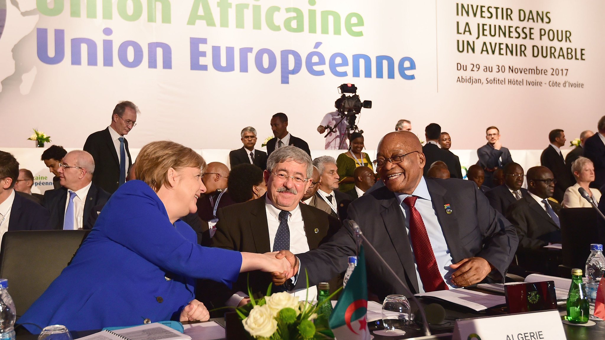 L'image montre la scène d'une conférence internationale où des dirigeants participent à un sommet. Au premier plan, on aperçoit un groupe de personnes, parmi lesquelles une femme, identifiée comme une figure politique importante, est en train de serrer la main d'un homme. Cette interaction symbolise le dialogue et la collaboration entre les nations. À l'arrière-plan, on distingue des bannières et des affiches portant le titre "Union Africaine, Union Européenne" ainsi que le slogan "Investir dans la jeunesse pour un avenir durable". La salle semble remplie de délégués, en costard et concentrés sur les discussions, ce qui donne une atmosphère formelle et engagée. Des éléments comme une table avec des drapeaux de différents pays ajoutent au cadre diplomatique de l'événement.