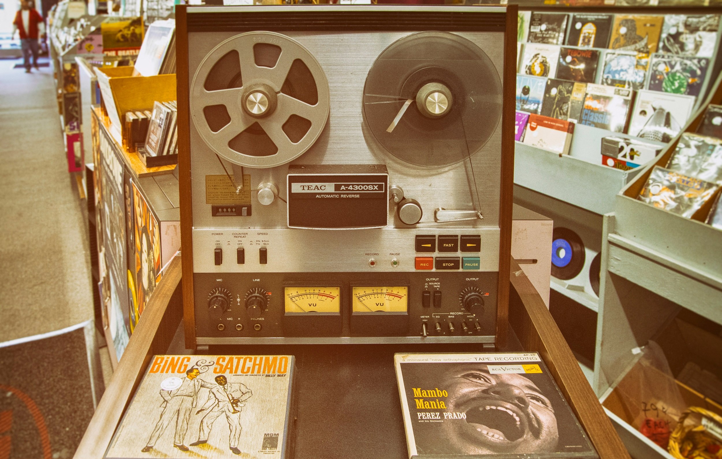 L'image montre un équipement audio vintage, un magnétophone à bande TEAC A-2300X, qui est posé sur un meuble en bois. Ce magnétophone a un design classique, avec un boîtier en métal argenté et des bobines de bande visibles sur le dessus. Deux compteurs analogiques sont situés en bas, indiquant les niveaux audio. À côté, on peut voir deux albums vinyles : l'un est intitulé "Bing & Satchmo", qui met en avant une collaboration entre le célèbre chanteur Bing Crosby et Louis Armstrong. L'autre album, "Mambo Mania" de Pérez Prado, suggère une atmosphère festive et rythmée. À l'arrière-plan, on aperçoit des étagères avec d'autres disques vinyles, créant une ambiance nostalgique et musicale.