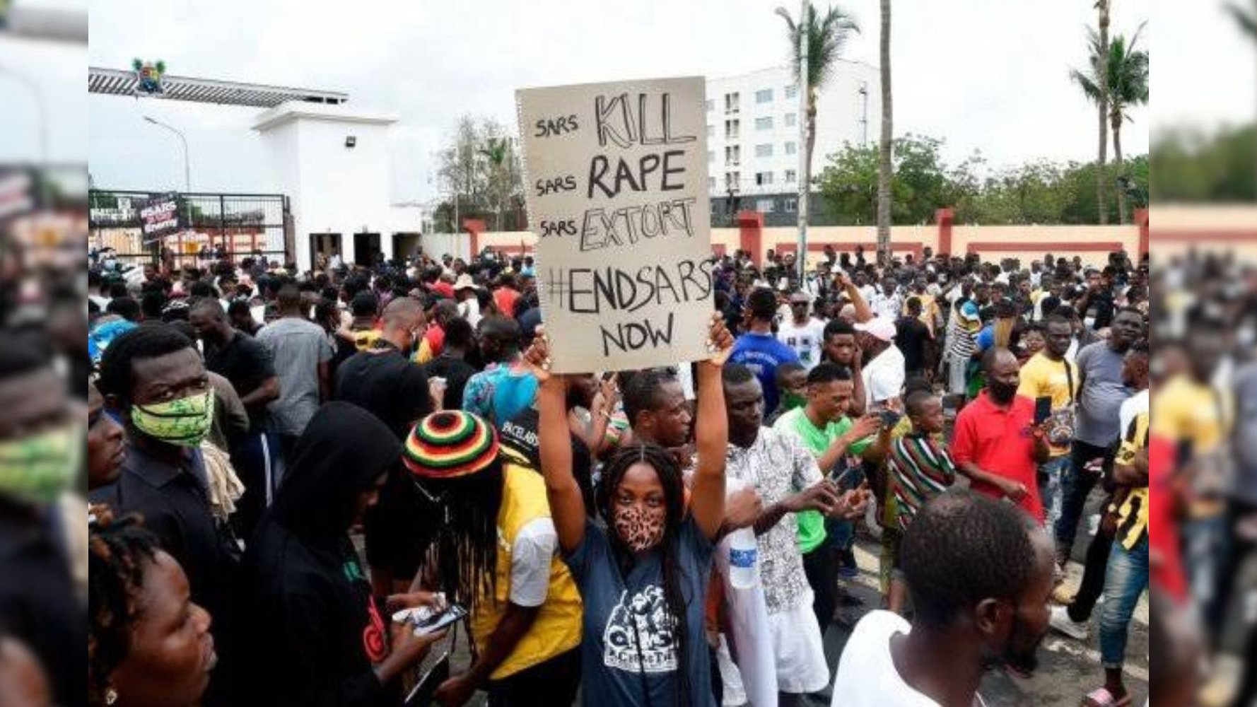 L'image montre une foule dense rassemblée pour une manifestation. Au centre, une personne tient une grande pancarte avec des inscriptions demandant la fin d'abus de la police. Les manifestants portent des vêtements variés, certains affichent des signes de solidarité, comme des masques. En arrière-plan, on aperçoit des bâtiments et des palmiers, ce qui donne une idée que l'événement se déroule dans une ville. L'atmosphère est chargée d'énergie collective, marquée par un sentiment de lutte et de revendication pour des droits.