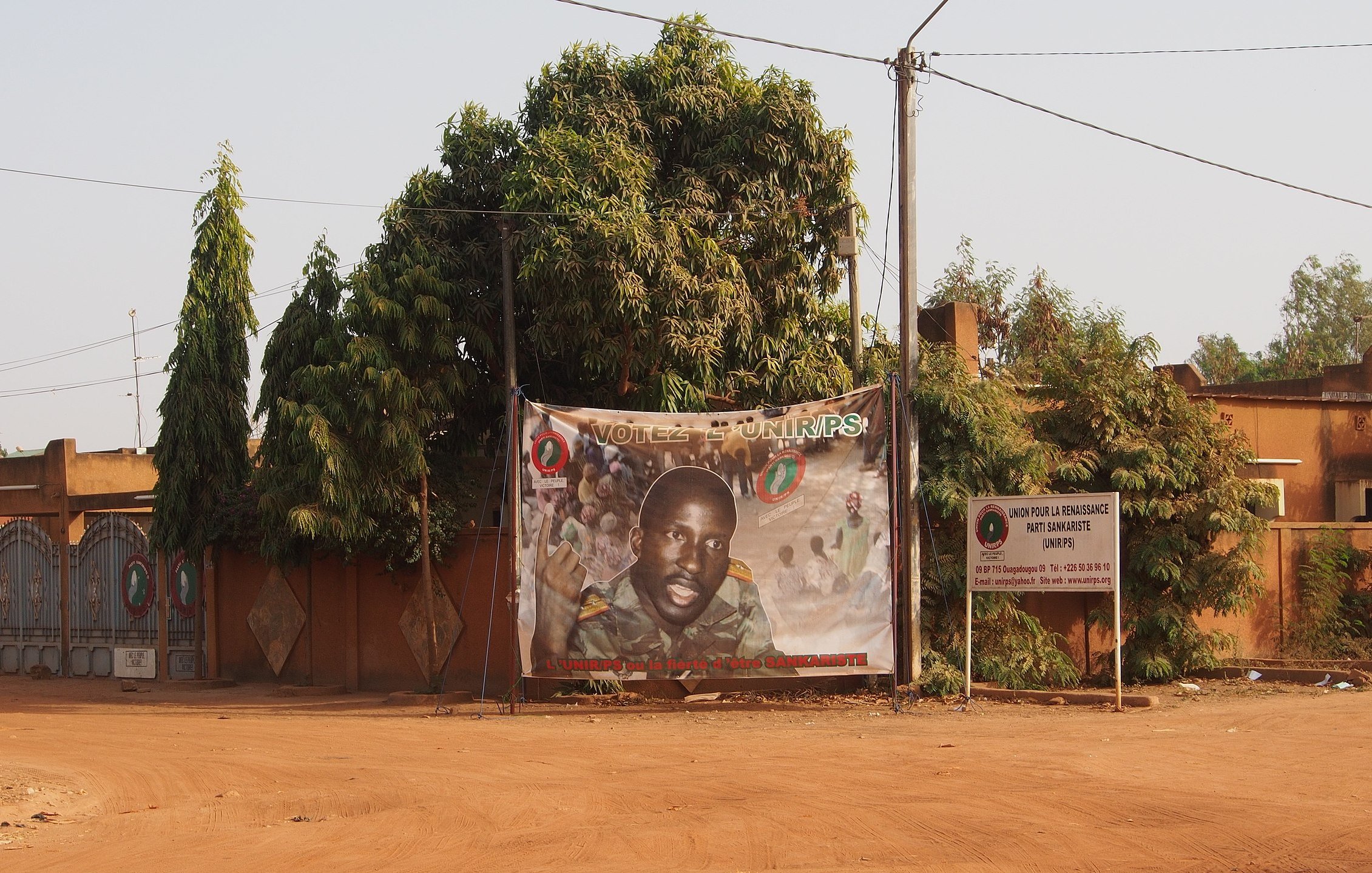 L'image présente une rue déserte d'un quartier, où le sol est recouvert de terre ocre. À gauche, un grand arbre luxuriant, avec des feuilles vertes, apporte une touche de nature. Au centre, une affiche enroulée sur un grillage présente le portrait d'un homme, probablement un candidat politique, souriant et regardant vers l'objectif. L'affiche est ornée de divers motifs colorés et de slogans. À droite, on peut distinguer un panneau d'information sur les élections. Les murs en arrière-plan sont peints dans des tons chauds, contribuant à l'atmosphère locale et authentique de l'endroit.
