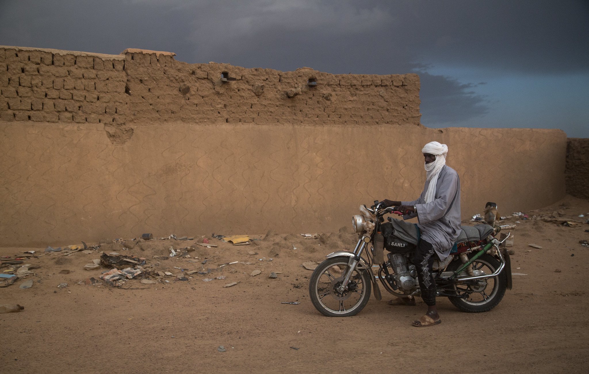 L'image montre un homme immobile à côté de sa moto dans un paysage désertique. Il porte un long vêtement traditionnel et un turban qui couvre sa tête, laissant seulement ses yeux visibles. Le sol est constitué de sable et de débris, tandis qu'en arrière-plan, on distingue un mur de terre crue, simple et robuste, typique des constructions dans cette région. Le ciel est nuageux, ajoutant une atmosphère dramatique à la scène. L'homme semble contemplatif, connectant ainsi l'élément humain avec l'environnement désertique qui l'entoure.