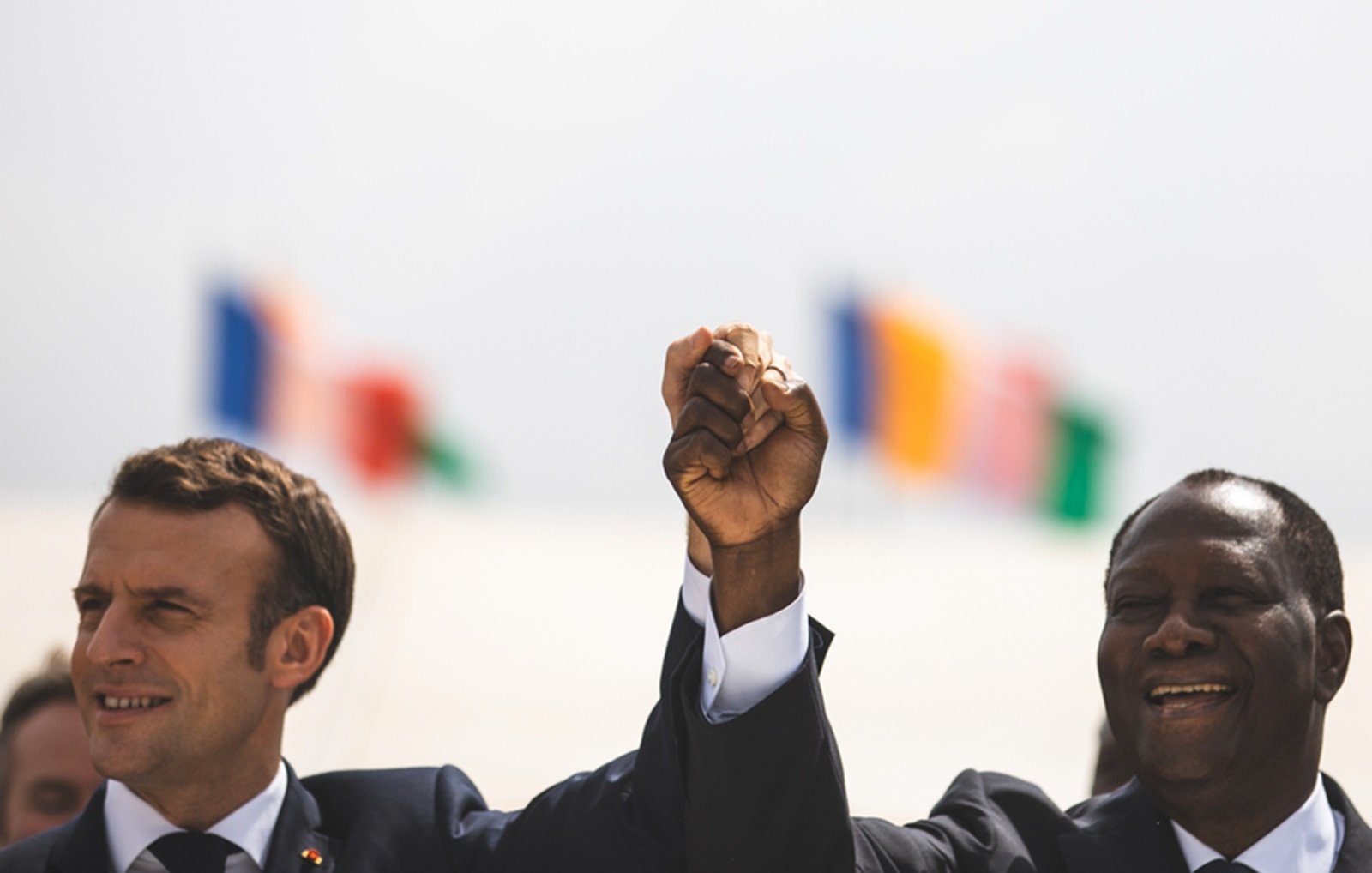 Imaginez une scène marquée par un moment de camaraderie et d'unité. Deux hommes se tiennent côte à côte, levant la main droite en l'air, leurs poings serrés, symbolisant la force et la solidarité. À gauche, on perçoit un homme avec une coupe de cheveux soignée, vêtu d'un costume sombre, tandis que l'homme à droite, également en costume, affiche un grand sourire chaleureux. En arrière-plan, plusieurs drapeaux flottent doucement au vent, représentant différentes nations. L'atmosphère est festive et pleine d'espoir.