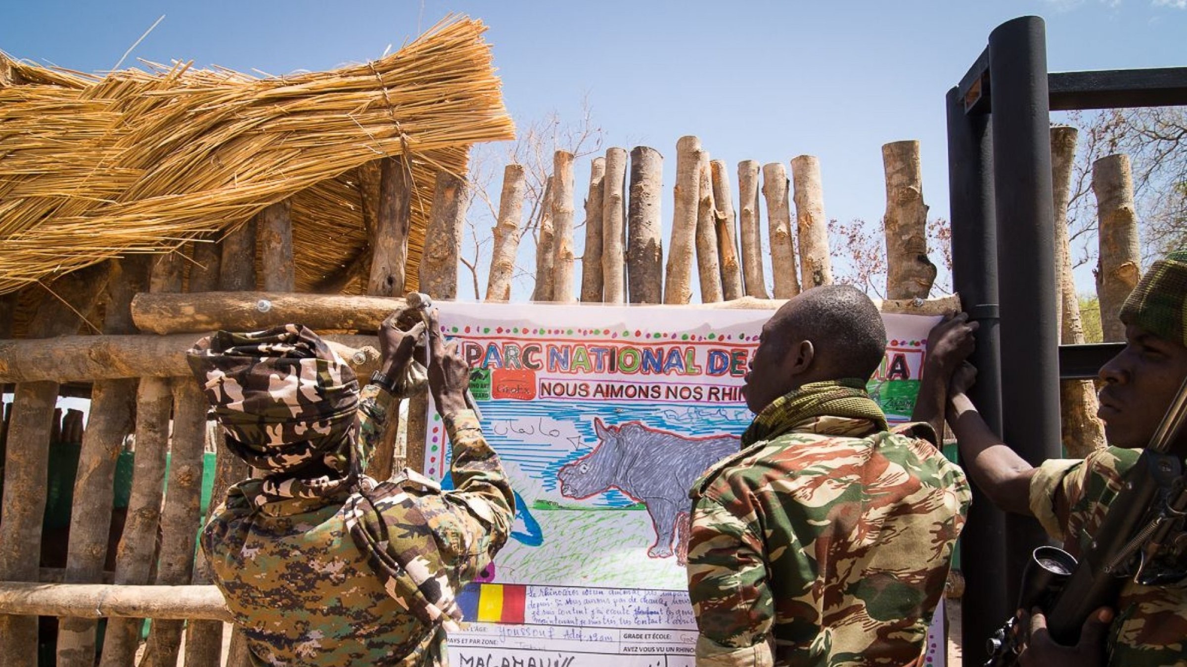 Sur l'image, nous voyons deux soldats en uniforme coloré, engagés dans une action collective. Ils sont en train d'accrocher une grande affiche sur un mur en bois fait de branches. L'affiche représente un rhinocéros, avec un dessin coloré et un message écrit en français qui exprime un engagement envers la protection de cette espèce. Le cadre est en plein air, avec un ciel bleu et quelques éléments naturels visibles. L'atmosphère dégage un sentiment de protection et d'éducation environnementale, soulignant l'importance de préserver la faune.