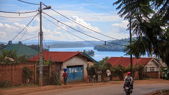 Voici une description de l'image : Vous vous trouvez sur une route située dans un paysage rural. À votre gauche, des maisons aux toits de tôle rouge et bleu se dressent, entourées de verdure luxuriante. Au loin, un lac scintille sous la lumière du soleil, reflétant les nuages blancs dans un ciel bleu. Certains habitants se déplacent, l'un d'eux portant une charge sur sa tête, tandis qu'un motard passe sur la route. Des poteaux électriques bordent la rue, ajoutant une touche de modernité à ce cadre naturel paisible. Le son lointain de l'eau et le chant des oiseaux peuvent également être présents dans cette ambiance tranquille.