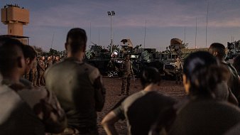 L'image montre un groupe de soldats rassemblés, attentifs, dans un environnement désertique au crépuscule. Ils sont partiellement de dos, ce qui met en avant leur tenue militaire. À l'arrière-plan, plusieurs véhicules militaires sont visibles, équipés d'armements. Le ciel est teinté de nuances douces, marquant la transition entre le jour et la nuit. L'atmosphère semble concentrée, suggérant une préparation ou un briefing important pour les soldats sur le terrain.