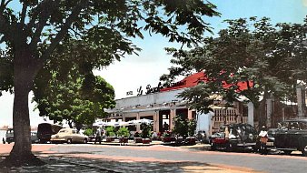 L'image montre une scène d'un restaurant, probablement située dans un quartier ensoleillé. À l'avant, on aperçoit une route bordée d'arbres dont les feuilles offrent un peu d'ombre. Le bâtiment principal, assez spacieux, possède un toit rouge et est marqué par l'inscription "Le Restaurant" en lettres blanches. Plusieurs voitures anciennes, de différentes couleurs, sont garées près de l'établissement. Des personnes flânent autour, créant une atmosphère de vie et de convivialité. En arrière-plan, le ciel est bleu, ce qui ajoute une touche de chaleur et de clarté à l'ensemble de l'image.