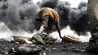L'image montre un environnement très sombre et pollué, avec d'épaisses colonnes de fumée noire s'élevant dans l'air. Au premier plan, un jeune homme est accroupi, en train de manipuler des débris. Il porte des vêtements simples, à moitié sales, et semble concentré sur ce qu'il fait. Son environnement est chaotique, avec des morceaux de métal, de la ferraille et des pneus éparpillés autour de lui. La scène dégage une atmosphère de difficulté et de lutte, illustrant les conditions de vie dans cet endroit touché par la pollution et la dégradation environnementale.