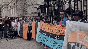 Dans cette image, un groupe de manifestants se rassemble devant un édifice imposant, probablement un bâtiment gouvernemental. Les personnes sont vêtues de vêtements variés, et certaines tiennent des pancartes. L'une des pancartes est colorée en orange, noir et bleu, et porte des inscriptions comme "Back to our Paradise" et "Forgotten Chagos". L'atmosphère est empreinte de détermination, avec des visages sérieux et engagés. Au fond, on peut voir des policiers surveillant la situation. L'environnement est urbain, avec des pavés au sol et des murs architecturaux ornés.