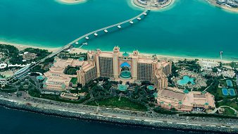 Cette image offre une vue aérienne d'un majestueux complexe hôtelier, entouré par des eaux turquoise. Le bâtiment principal, qui semble imposant, est décoré de nuances de beige et d'or, avec une architecture inspirée des contes arabes. Des jardins luxuriants entourent l'hôtel, agrémentés de piscines scintillantes et de palmiers. À proximité, on peut voir un chemin qui s'étend au-dessus de l'eau, reliant le terre-plein à d'autres structures. L'ensemble dégage une atmosphère luxueuse et tropicale, évoquant des vacances chaleureuses.