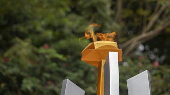 L'image représente une flamme allumée sur un socle jaune, qui surplombe plusieurs structures géométriques grises. La flamme danse avec vivacité, créant une ambiance dynamique, entourée d'un fond verdoyant flou, suggérant la présence d'arbres et de végétation. Cette scène évoque un symbole de passion et de célébration, souvent associé à des événements prestigieux comme les Jeux Olympiques. L'ensemble dégage une impression de force et de détermination.