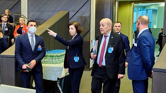 Dans cette scène, un groupe de personnes se déplace à l'intérieur d'un bâtiment moderne, probablement lors d'une réunion ou d'un événement officiel. Au premier plan, une femme, vêtue d'un costume professionnel et portant un document à la main, pointe dans une direction, semblant donner des indications. À ses côtés, un homme portant un masque de protection écoute attentivement, tandis qu'un autre homme, en costume noir, marche à ses côtés, apparemment engagé dans une conversation. En arrière-plan, d'autres participants sont visibles, suggérant une atmosphère animée et collaborative. Des bouteilles d'eau sont disposées sur une table à proximité, signalant la durée de l'événement. Le décor est contemporain, avec des murs en bois et des éléments de design épuré.