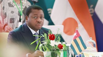 L'image montre un homme assis à une table lors d'un événement officiel. Il porte un costume sombre et une cravate, et son expression semble sérieuse et attentive. Sur la table, il y a un petit vase avec des fleurs colorées, ajoutant une touche de vie à la scène. À côté de lui, on voit un drapeau qui représente le Togo. En arrière-plan, des drapeaux d'autres pays sont également visibles, indiquant un cadre international. L'atmosphère semble formelle et concentrée.