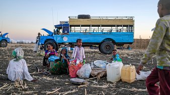 L'image montre un groupe de personnes assises sur un sol de terre battue, entourées de paniers et de contenants. Au centre, des femmes et des enfants semblent converser, tandis qu'un enfant se tient à droite, s'approchant. En arrière-plan, on peut voir un bus bleu immobile, avec un ciel dégradé qui passe des teintes orangées aux bleues, suggérant un crépuscule. Le terrain est jonché de restes de culture, ajoutant un contexte rural à la scène. L'atmosphère évoque une ambiance de rassemblement communautaire dans un environnement de travail.