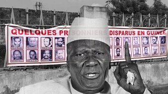 L'image montre un homme d'âge mûr, vêtu d'une tenue traditionnelle et portant un chapeau. Il lève un doigt en signe d'alerte ou de demande d'attention. En arrière-plan, on peut voir une grande banderole accrochée sur un mur. Cette banderole affiche des portraits de plusieurs personnes, accompagnés d'un texte qui fait référence à des prisonniers disparus en Guinée. L'ambiance de l'image évoque un appel à ne pas oublier ces disparitions et à sensibiliser le public à cette problématique.