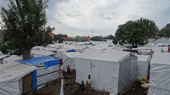 Cette image montre un paysage d'un camp de réfugiés, avec de nombreuses tentes blanches disposées de manière un peu désordonnée. Les tentes sont recouvertes d'une toile claire qui les protège des intempéries. On aperçoit quelques enfants jouant au milieu des tentes. Au loin, des arbres abondants ajoutent une touche de verdure, et on peut distinguer quelques structures plus solides. Le ciel est nuageux, suggérant qu'il pourrait pleuvoir, ce qui souligne la vulnérabilité du cadre. L'ambiance générale semble à la fois animée et calme, tandis que le sol est irrégulier, avec des pierres et des zones boueuses.