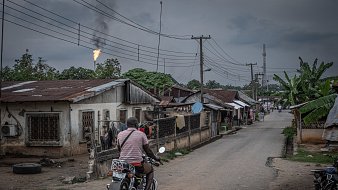 L'image présente une scène urbaine dans un quartier qui semble rural. Une route centrale est bordée de maisons modestes, souvent en tôle, avec des fenêtres grillagées. À gauche, on apperçoit un homme sur une moto, portant un t-shirt rayé, qui se dirige vers l'avant, ajoutant une touche de mouvement à la scène. En arrière-plan, des poteaux électriques sont visibles, ainsi qu'un tuyau de gaz qui rejette une flamme, indiquant peut-être une installation industrielle à proximité. Le ciel est couvert, avec des nuages sombres, créant une atmosphère légèrement mélancolique. À droite, des plantations de bananiers ajoutent une touche de verdure. L'ensemble dépeint une réalité de vie quotidienne dans un environnement à la fois simple et intéressant.