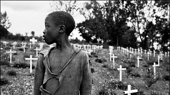L'image présente un jeune garçon qui se tient dans un cimetière parsemé de croix blanches, symbolisant des tombes. Son regard est tourné vers la gauche, et on peut percevoir une expression de réflexion ou de tristesse sur son visage. Il porte un vêtement déchiré, soulignant une certaine vulnérabilité ou pauvreté. En arrière-plan, le paysage est entouré d'arbres, créant une atmosphère de mélancolie. Les croix blanches, disposées sur la pente, évoquent la mémoire et le souvenir de ceux qui ont perdu la vie.