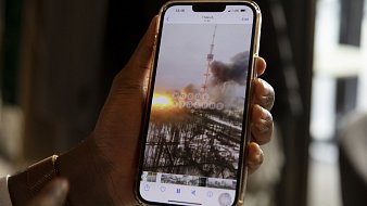 L'image montre une main tenant un smartphone dans un environnement intérieur. Sur l'écran du téléphone, on peut voir une photo qui semble représenter un paysage urbain, où une tour se dresse au milieu de colonnes de fumée et des nuages sombres. À l'arrière-plan, un coucher de soleil offre des teintes orange et jaune qui contrastent avec la grisaille de la scène. Les détails flous et éthérés suggèrent une atmosphère dramatique, peut-être liée à un événement perturbant. La main qui tient le téléphone est d'une peau foncée et porte un bracelet lumineux, ajoutant une touche personnelle à l'image.