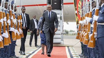 L'image montre un moment officiel sur un tapis rouge, où un homme en costume, probablement un dignitaire, marche avec assurance. Il est entouré de garde d'honneur en tenue formelle, tenant des fusils. À l'arrière-plan, on aperçoit une grande échelle d'avion et d'autres personnes en costume. L'atmosphère dégage un sentiment de cérémonie et de respect, soulignée par la disposition soignée des militaires et le faste du décor.