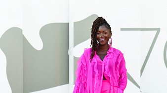 L'image montre une femme debout sur un fond minimaliste. Elle porte une tenue éclatante de couleur rose vif, composée d'une blouse légère aux manches froncées et d'une jupe assortie. Son sourire est radieux et elle affichent une attitude pleine de confiance. Ses cheveux sont coiffés en dreadlocks, et elle porte de grandes boucles d'oreilles qui ajoutent une touche élégante à son look. Le fond est composé de formes géométriques en tons neutres, mettant en valeur la vivacité de sa tenue.