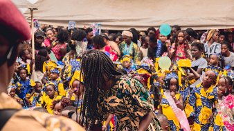 L'image montre une scène vibrante et animée d'un événement en plein air, probablement une célébration ou un festival. Au premier plan, on voit une femme portant une robe aux motifs colorés qui se penche, en train de distribuer quelque chose. Derrière elle, un grand groupe de filles, habillées de robes jaunes ornées de motifs bleus, se tient avec des sourires sur le visage. Leur ambiance semble joyeuse et festive. La foule est dense, avec de nombreuses personnes, y compris des femmes et des enfants, qui interagissent et profitent de l'événement. Des éléments décoratifs, comme des ballons ou des banderoles, pourraient ajouter une touche festive à l'environnement. L'atmosphère globale dégage une sensation de communauté et de célébration.