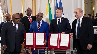L'image montre un groupe de personnes lors d'une cérémonie officielle. Au centre, deux hommes tiennent des documents signés, symbolisant un accord ou un partenariat. L'un d'eux porte un costume bleu avec une cravate à motifs, tandis que l'autre est vêtu d'un costume sombre avec une cravate noir. En arrière-plan, on remarque des drapeaux, dont ceux de la France et d'autres pays africains. Les autres personnes en arrière-plan observant la scène portent également des costumes. L'ambiance générale est solennelle, mettant en avant un moment important de collaboration entre les nations.