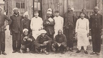 L'image montre un groupe de soldats sénégalais convalescents rassemblés devant un bâtiment en bois, vraisemblablement un hôpital de campagne. Il y a treize personnes, dont des hommes en uniforme militaire et des membres du personnel médical. Les soldats portent des uniformes sombres et certains d'entre eux ont des casquettes ou des bonnets. Le personnel médical, d'autre part, porte des blouses blanches, indiquant leur rôle. Tous affichent des expressions amicales, certains sourient tandis que d'autres prennent des poses plus sérieuses. L'arrière-plan montre des murs en bois, donnant une atmosphère de campagne ou de mobilisation militaire. Cette scène capture un moment de camaraderie et de résilience au sein des soldats pendant une période difficile.