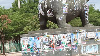 L'image montre une grande statue d'éléphant, probablement en bronze ou en métal, se tenant sur un socle en béton. La statue est recouverte de nombreuses affiches publicitaires et politiques, dont certaines sont déchirées et se détachent. Ces affiches présentent des visages et des textes, indiquant des campagnes électorales, des événements ou des promotions. Le socle en béton est partiellement peint en rouge et en gris, ajoutant un contraste à l'image. À l'arrière-plan, on aperçoit des arbres verdoyants et d'autres éléments de la rue, ce qui suggère une ambiance vivante et urbaine. Les couleurs sont variées et l'aspect général est un mélange de textures et de visuels, témoignant d'une interaction entre la nature et l'urbanisme.
