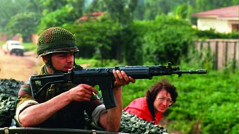 L'image montre un soldat, assis dans un véhicule militaire,tenant un fusil vers l'avant. Il est vêtu d'un uniforme camouflage et porte un casque en treillis. Son visage exprime la concentration et la vigilance, tandis qu'il regarde attentivement son environnement. En arrière-plan, une femme à lunettes observe la scène, semblant préoccupée. La verdure luxuriante entoure le cadre, créant un contraste avec la terre battue sur laquelle se trouve le véhicule. L'atmosphère de l'image est chargée de tension, suggérant un contexte de vigilance et de protection.