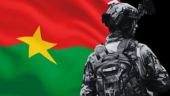 L'image montre un soldat de dos, vêtu d'un uniforme de camouflage, avec un sac à dos. En arrière-plan, se trouve le drapeau du Burkina Faso, qui est composé de bandes horizontales rouges et vertes avec une étoile jaune au centre. Le soldat est positionné de manière à mettre en valeur le drapeau, symbolisant probablement la fierté nationale ou le service militaire.