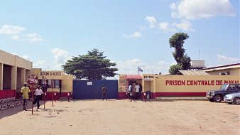 L'image montre l'entrée de la Prison Centrale de Makala. Devant le bâtiment, on voit une grande porte bleue. À droite, une cabine d'accès avec des petits guichets, probablement pour les visiteurs. Des personnes sont sur le point d'entrer ou de sortir. L'environnement est désertique, avec du sable au sol. Quelques voitures sont garées à droite, et un grand arbre se dresse à proximité. Le ciel est clair, avec quelques nuages. Des inscriptions sont visibles sur les murs, notamment le nom de la prison.