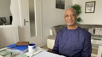 L'image montre un homme assis à une table chez lui. Il porte une tenue traditionnelle de couleur sombre et affiche un sourire chaleureux. La table est en verre et est ornée de divers objets, dont un pot de fleurs vert, un carnet ouvert avec des notes, et un stylo. À côté du carnet, il y a une tasse bleue sur la table. En arrière-plan, on peut voir un canapé clair et des décorations murales. La lumière naturelle entre par une fenêtre, créant une ambiance lumineuse et accueillante.