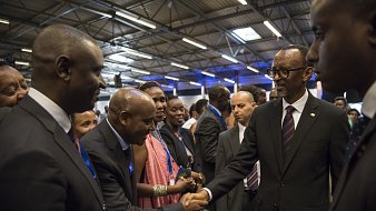 Dans cette image, nous voyons un groupe de personnes rassemblées dans un espace intérieur, probablement lors d'un événement officiel ou d'une conférence. Parmi eux se trouve un homme en costume noir avec une cravate, qui semble être un dirigeant ou une personne importante. Il tend la main pour saluer un autre homme, également en costume. Les autres personnes présentes, principalement habillées formellement, regardent avec intérêt. L'atmosphère est conviviale et respectueuse, avec des sourires et des échanges amicaux. Les murs sont décorés de manière simple, et un éclairage doux illumine la scène.