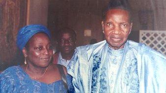 L'image montre trois personnes. À l'avant, un homme et une femme sont vêtus de costumes traditionnels, probablement en tissus d'importance culturelle. L'homme porte un chapeau et a un regard sérieux, tandis que la femme a un sourire et semble détendue. En arrière-plan, un troisième homme est visible, bien qu'il soit moins en avant-plan. L'ambiance de l'image semble formelle.