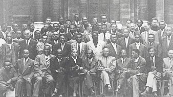 L'image présente un groupe de plusieurs hommes, probablement des leaders et des intellectuels, rassemblés pour une photographie de groupe. Ils sont vêtus de costumes formels, avec une variété de poses : certains sont assis, d'autres debout. L'ambiance semble chaleureuse et conviviale. Au centre, une femme se distingue, assise avec un sourire. En arrière-plan, on aperçoit un bâtiment historique, suggérant peut-être un événement ou une rencontre importante. L'image est en noir et blanc, ce qui lui donne un aspect intemporel.