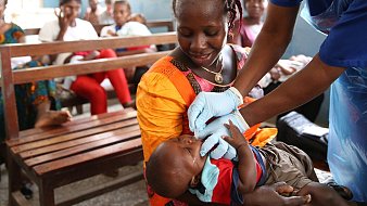 Dans cette image, nous voyons une scène dans une salle d'attente où plusieurs personnes sont assises sur des bancs en bois. Au premier plan, une mère, vêtue d'une robe orange, tient son bébé. Elle semble attentive et douce. Un professionnel de la santé, portant des gants bleus, est en train de donner un soin ou un médicament au bébé. Le bébé, habillé d'un t-shirt bleu et d'un short, a une expression calme. À l'arrière-plan, d'autres femmes et enfants attendent, créant une atmosphère de communauté et de soutien. Les murs sont peints en bleu, ajoutant une ambiance sereine à la scène.