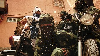 L'image montre un groupe de personnes habillées de manière distinctive, portant des vêtements camouflés, ainsi que des masques et des foulards ornés de paillettes ou de motifs. Ils se trouvent dans une rue d'une vieille ville, probablement dans un environnement urbain. À côté d'eux, deux motos ajoutent un élément de modernité à la scène. Leurs poses évoquent une certaine solidarité et un esprit de camaraderie. L'éclairage est naturel, suggérant un moment de la journée typique dans une ville animée. Les textures des vêtements contrastent avec le pavé de la rue, créant une ambiance unique.