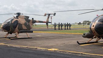 L'image montre une scène aérienne sur un terrain d'aviation. Deux hélicoptères, de couleur beige avec des motifs camouflage, sont stationnés sur le tarmac. À l'arrière-plan, un groupe de soldats en uniforme se dirige en formation vers un point de destination, créant une atmosphère dynamique et militaire. Le ciel est nuageux, ajoutant une ambiance dramatique à la scène. Le paysage autour est dégagé, avec des bâtiments épars visibles au loin.