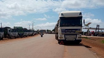 L'image montre une route large et droite, où une file de camions est stationnée sur le côté. Les camions, de différentes tailles et couleurs, sont alignés de manière ordonnée. Le ciel est clair avec quelques nuages blancs qui flottent. On peut apercevoir des poteaux électriques le long de la route, et au loin, il y a des bâtiments, suggérant un environnement urbain en arrière-plan. À proximité, un motocycliste passe, ajoutant un peu de dynamisme à la scène calme. L'ambiance générale dégage une impression de circulation routière et d'activités commerciales.
