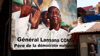 L'image représente une grande affiche murale d'un homme en train de saluer, avec un sourire bienveillant. Il porte une tenue traditionnelle, incluant un chapeau blanc et une chemise claire. En arrière-plan, on peut voir des couleurs vives qui semblent évoquer une atmosphère festive ou patriotique. Au bas de l'affiche, le texte indique « Général Lansana Conté, Père de la démocratie multipartite », soulignant ainsi son rôle important dans l'histoire politique. Les lignes et les formes des éléments de l'affiche créent un contraste qui attire le regard, suggérant un message de respect et d'honneur envers cette figure politique.