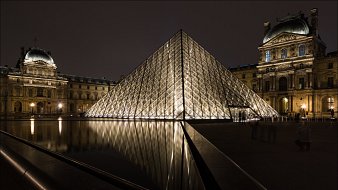 L'image montre le célèbre musée du Louvre à Paris, avec sa pyramide en verre illuminée au centre de la scène. La pyramide, qui est moderne et de forme géométrique, se dresse majestueusement au-dessus d'un bassin d'eau calme qui en reflète la lumière. À gauche, on aperçoit l'architecture historique du musée, avec ses pierres anciennes et des détails raffinés. La scène est plongée dans une ambiance nocturne, où les lumières chatoyantes se mêlent à l'obscurité, créant un contraste saisissant entre le moderne et le classique. Le reflet dans l'eau ajoute une dimension poétique à l'ensemble.