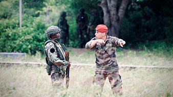 L'image montre deux militaires en discussion dans un environnement extérieur. À gauche, un soldat vêtu d'un uniforme camouflage, portant un casque et tenant un fusil. À droite, un autre militaire, qui porte une tenue camouflage et un béret rouge. Il semble gesticuler en indiquant une direction. En arrière-plan, on aperçoit d'autres soldats flous, ce qui suggère qu'ils sont dans une zone d'entraînement ou opérationnelle, entourée de végétation et d'arbres. L'ambiance est sérieuse et dénote une situation de coordination ou de briefing entre les membres de l'armée.