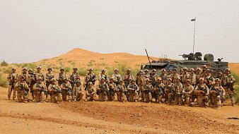 L'image montre un groupe de soldats rassemblés dans un paysage désertique. Au premier plan, on peut voir une trentaine de militaires, tous vêtus de tenues camouflées dans des tons de sable et de marron. Ils se tiennent debout ou accroupis, posant avec des expressions sérieuses. À l'arrière, un véhicule militaire, probablement un blindé, est stationné et porte un drapeau sur son toit. Le fond est marqué par des dunes de sable orangé, typiques des déserts, sous un ciel clair. Cette scène évoque un contexte militaire et une atmosphère de camaraderie entre les soldats.
